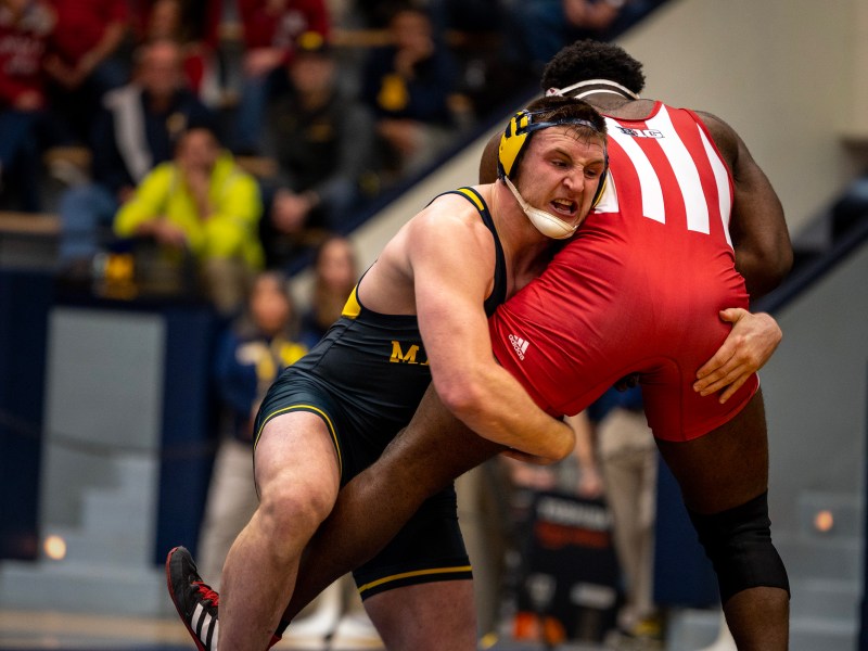 A Michigan wrestler grabs his opponent by the leg in an attempt to take him down as spectators watch on.