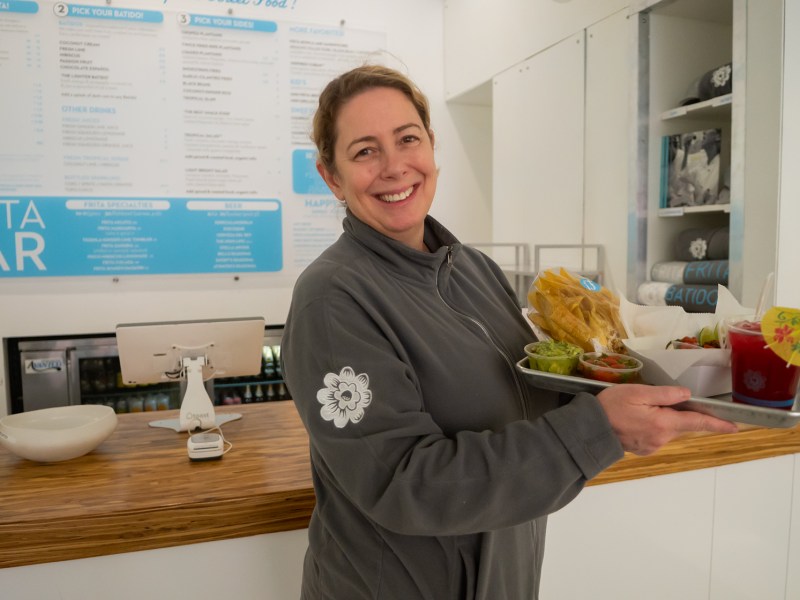 Eve Arnoff, the owner of Frita Batidos, holds up a plate with chips, salsa, guac and a batido. She is wearing a gray sweatshirt and behind her is the menu and a brown table with the checkout station.