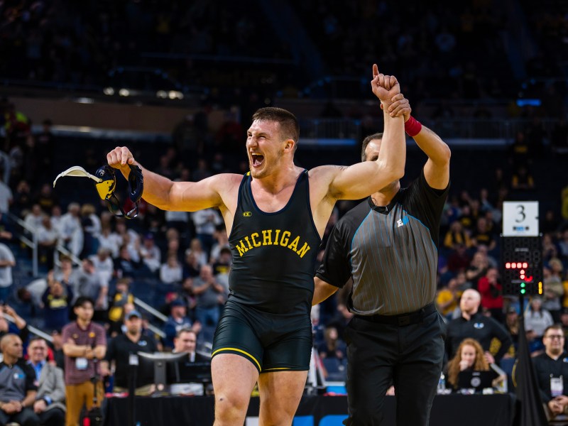 A Michigan wrestler excitedly yells while holding his headgear in one hand and the ref holds up his other hand to signal a victory. The wrestler holds up one finger with his hand and behind him the crowd cheers.