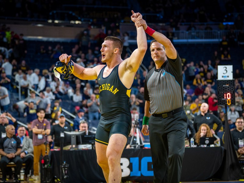 The referee declares the Michigan wrestler the winner of the match, holding the wrestler’s hand up in the air. The wrestler wears a navy and maize unitard and has an excited look on his face. A crowd of fans can be seen in the background.
