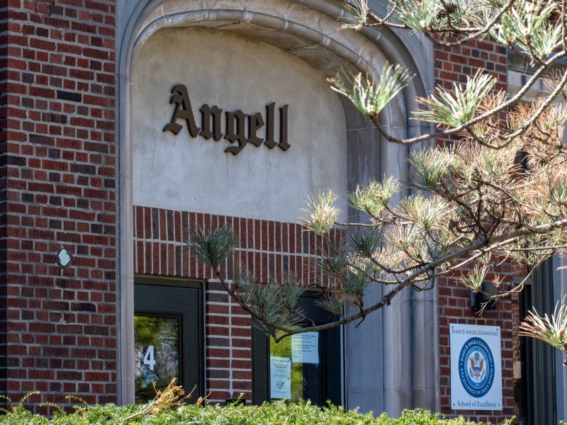 Sunlight shines on the branches of a pine tree in the foreground of the photo. In the background, a wooden sign that reads “Angell” in a cement arch hangs above a brick doorway. On the bottom right, there is a sign hanging that designates the building as a “School of Excellence.”