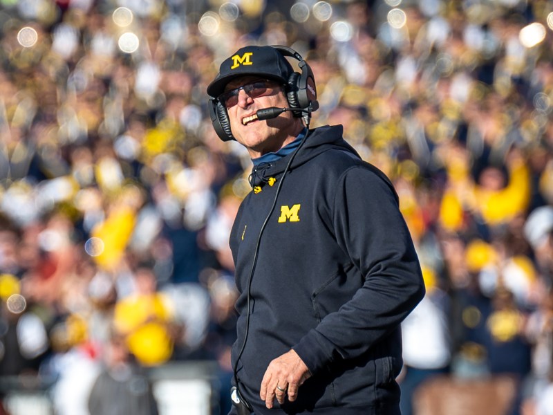 Jim Harbaugh stands on the field wearing a headset. He looks into the distance.