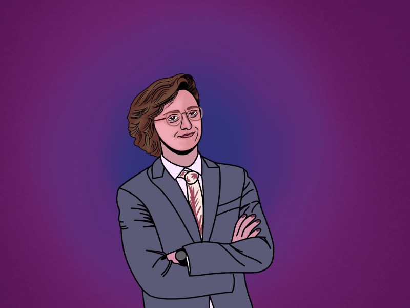 Brian David Gilbert in a suit standing with his arms crossed in front of a purplish background.