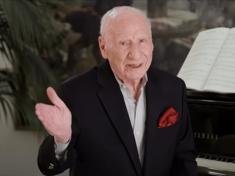 Comedian Mel Brooks smiling and gesturing to the camera while talking