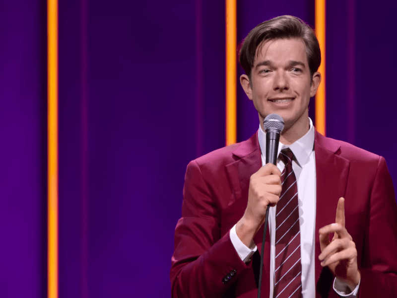 John Mulaney holding a microphone in a burgundy suit smiling while holding up one finger.