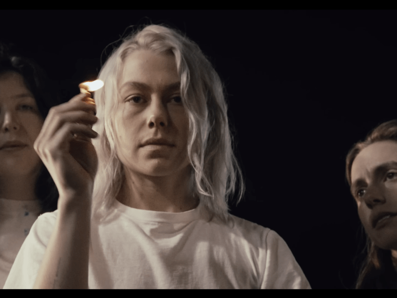 In a black void, Phoebe Bridgers holds a match in a white shirt while the other members of boygenius Lucy Dacus and Julien Baker look on in similar dress.